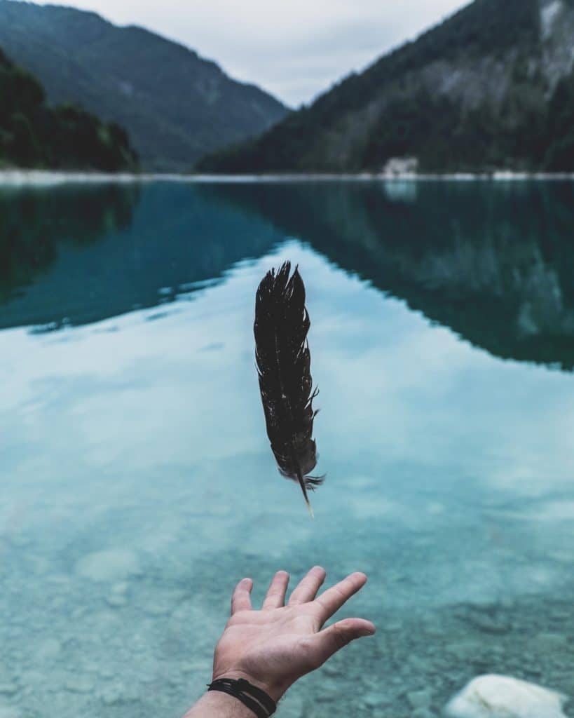 an open palm hand beneath a black feather with a calm water and 2 high mountains in the background 