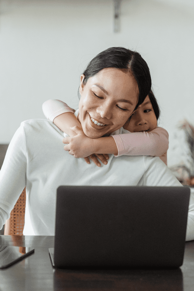 happy Asian mother working from home and little daughter hugging mom from behind