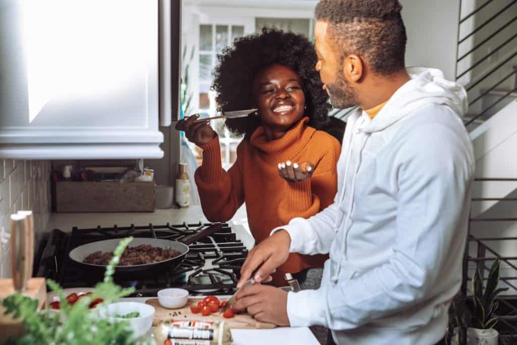a black woman wearing an orange sweatshirt, cooking and facing a black man wearing a white sweatshirt cooking together