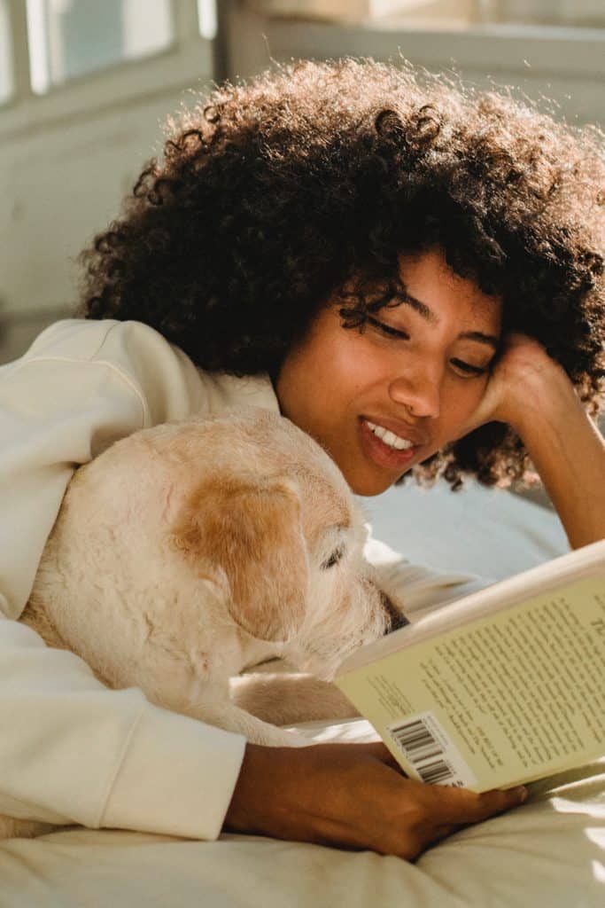 a black woman wearing a white top, lying in bed while hugging a dog and reading a book