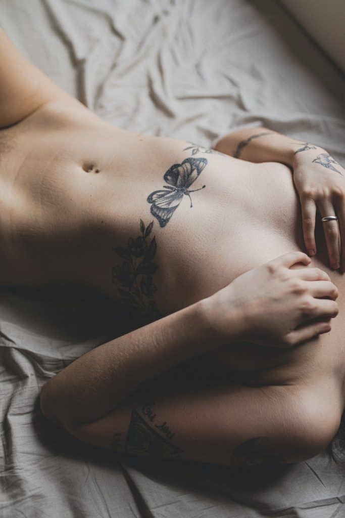 a naked woman with a chest tattoo lying in bed holding and covering her breast