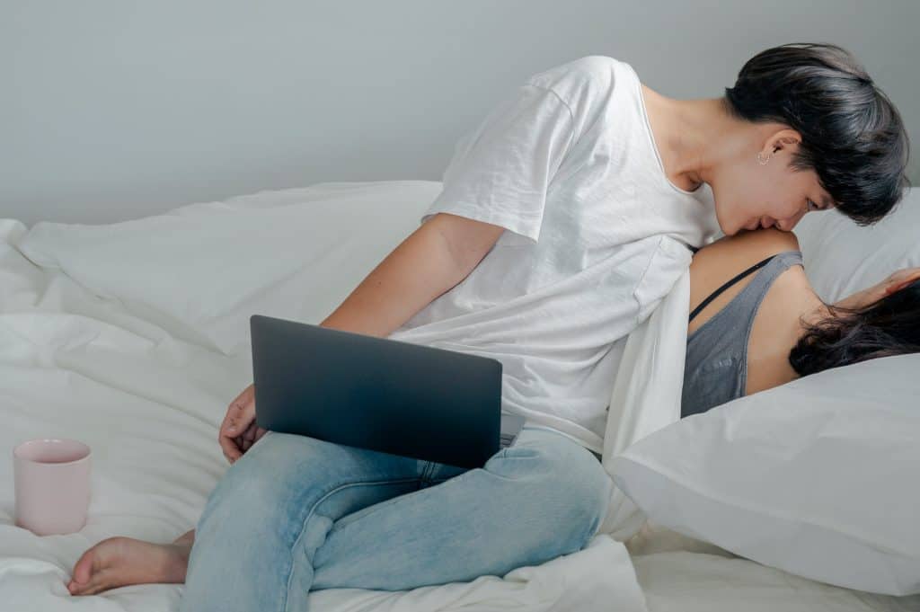 woman in white shirt with a computer on her lap, kissing a woman behind her lying in the couch
